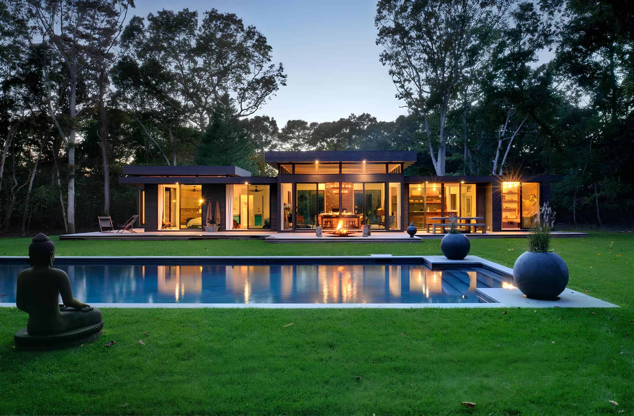 Modern contemporary house with large glass windows and outdoor pool, surrounded by lush greenery and tall trees, during sunset.