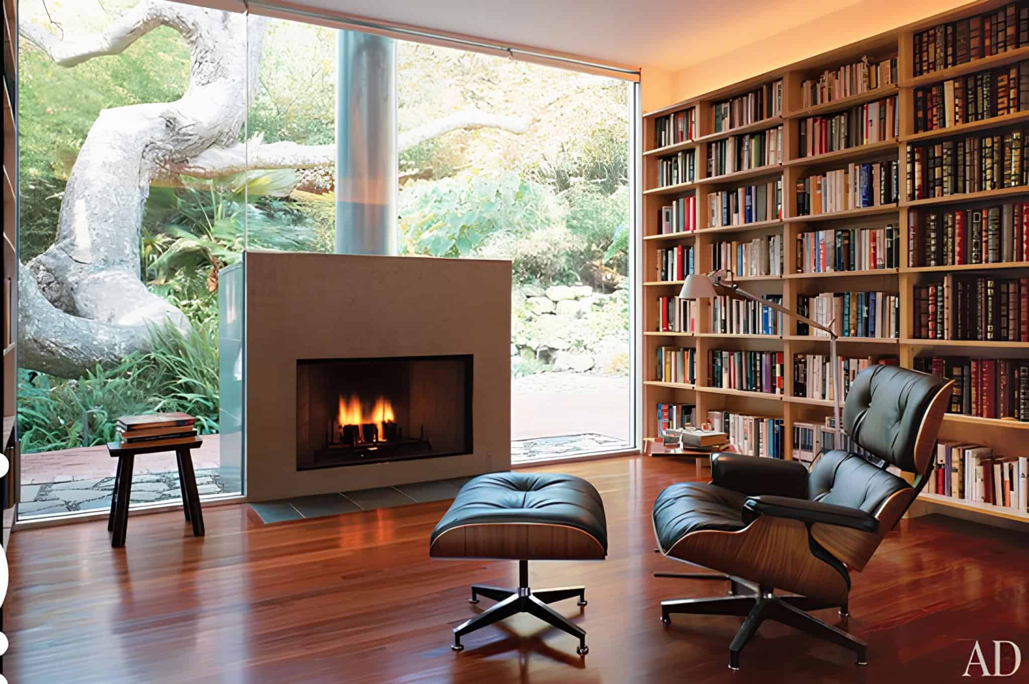 Cozy modern home library with large floor-to-ceiling window and indoor fireplace, furnished with a black leather lounge chair, wooden bookshelf filled with books, and a view of lush garden outside.