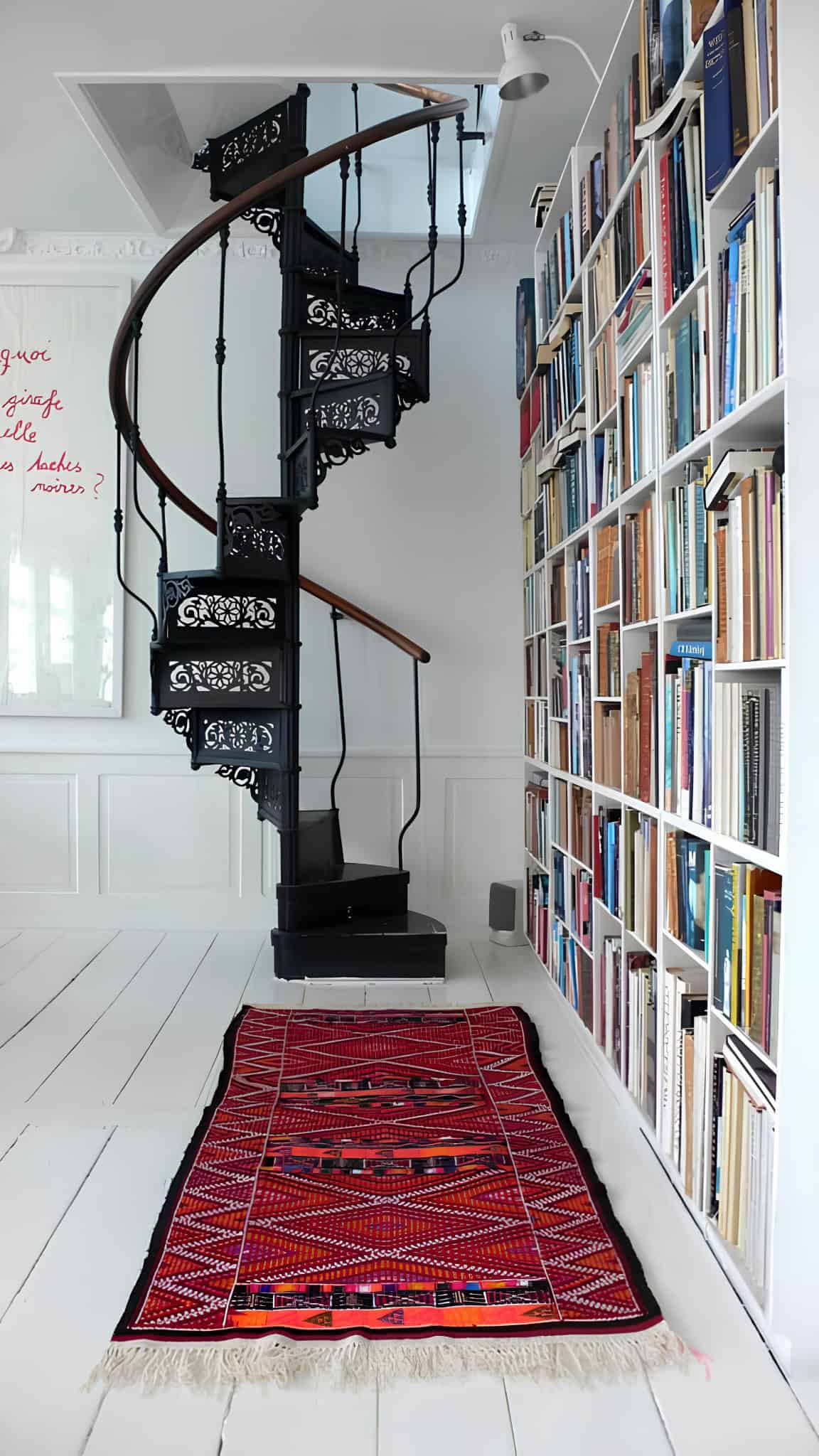 Narrow home library corner with black spiral staircase, tall book wall and red rug