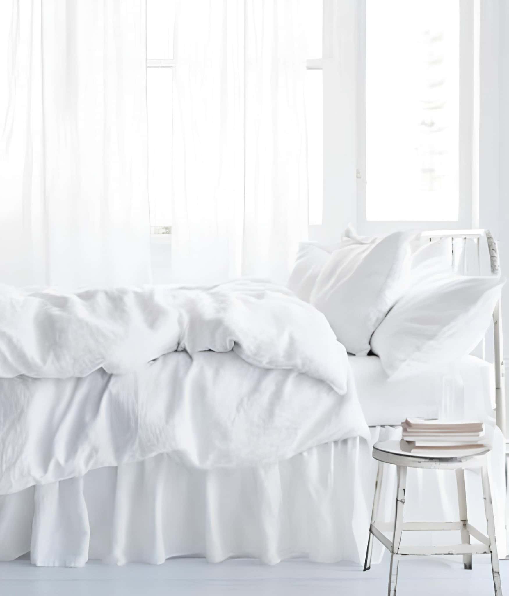 Minimalist White Bedroom with Sheer Curtains All-white bedroom with fluffy bedding, sheer curtains, and rustic stool