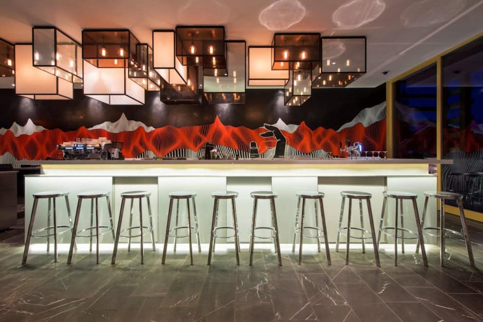 Contemporary bar design featuring illuminated white countertop, modern industrial stools, artistic mountain mural, and geometric ceiling light fixtures, perfect for stylish urban nightlife settings.