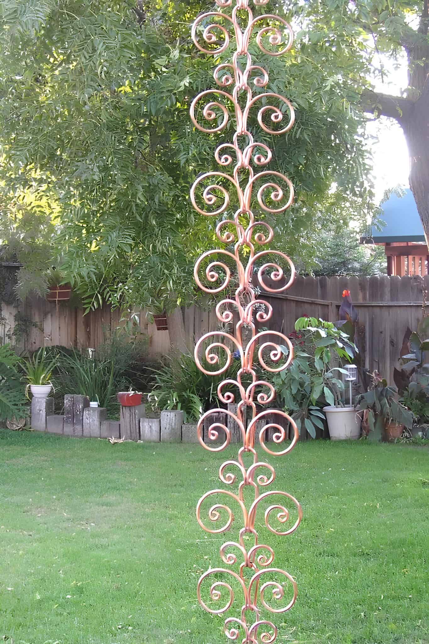 Elegant copper scrollwork rain chain hanging in a lush backyard garden