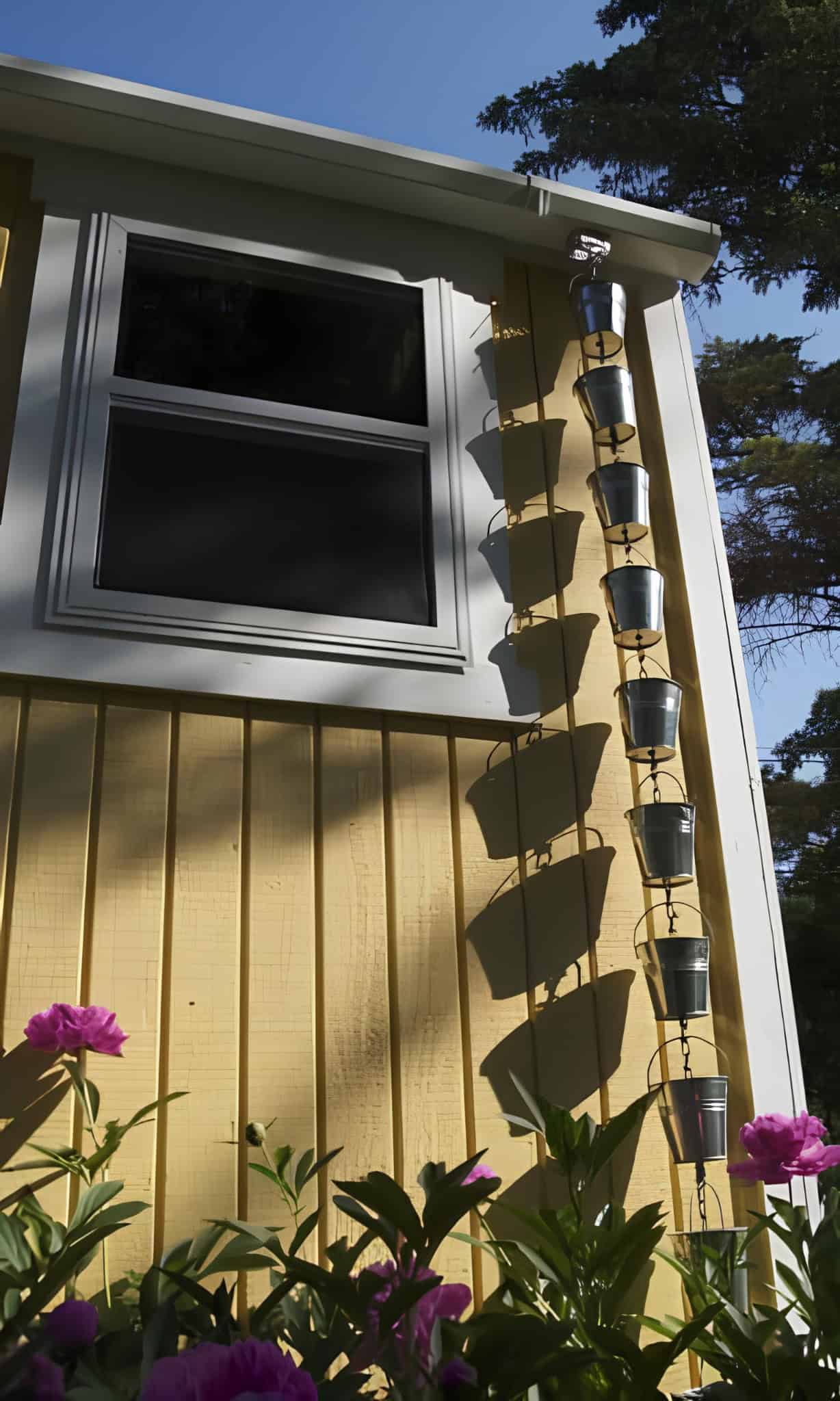 Metal bucket rain chain hanging from the gutter of a yellow house with flowers