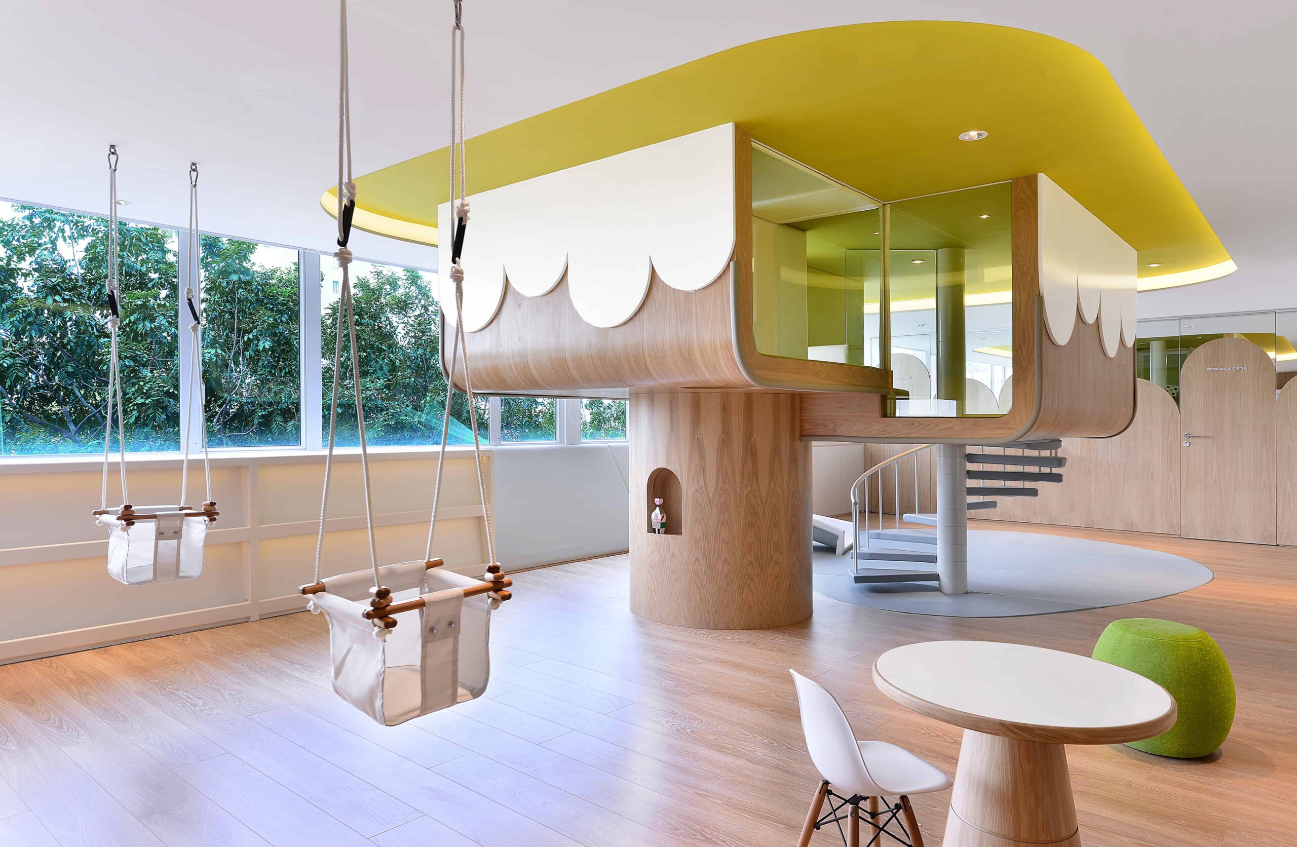 Modern indoor playground with treehouse design, swings, and playful furniture in a bright, spacious nursery or children's playroom. Natural light and contemporary decor create a welcoming environment.