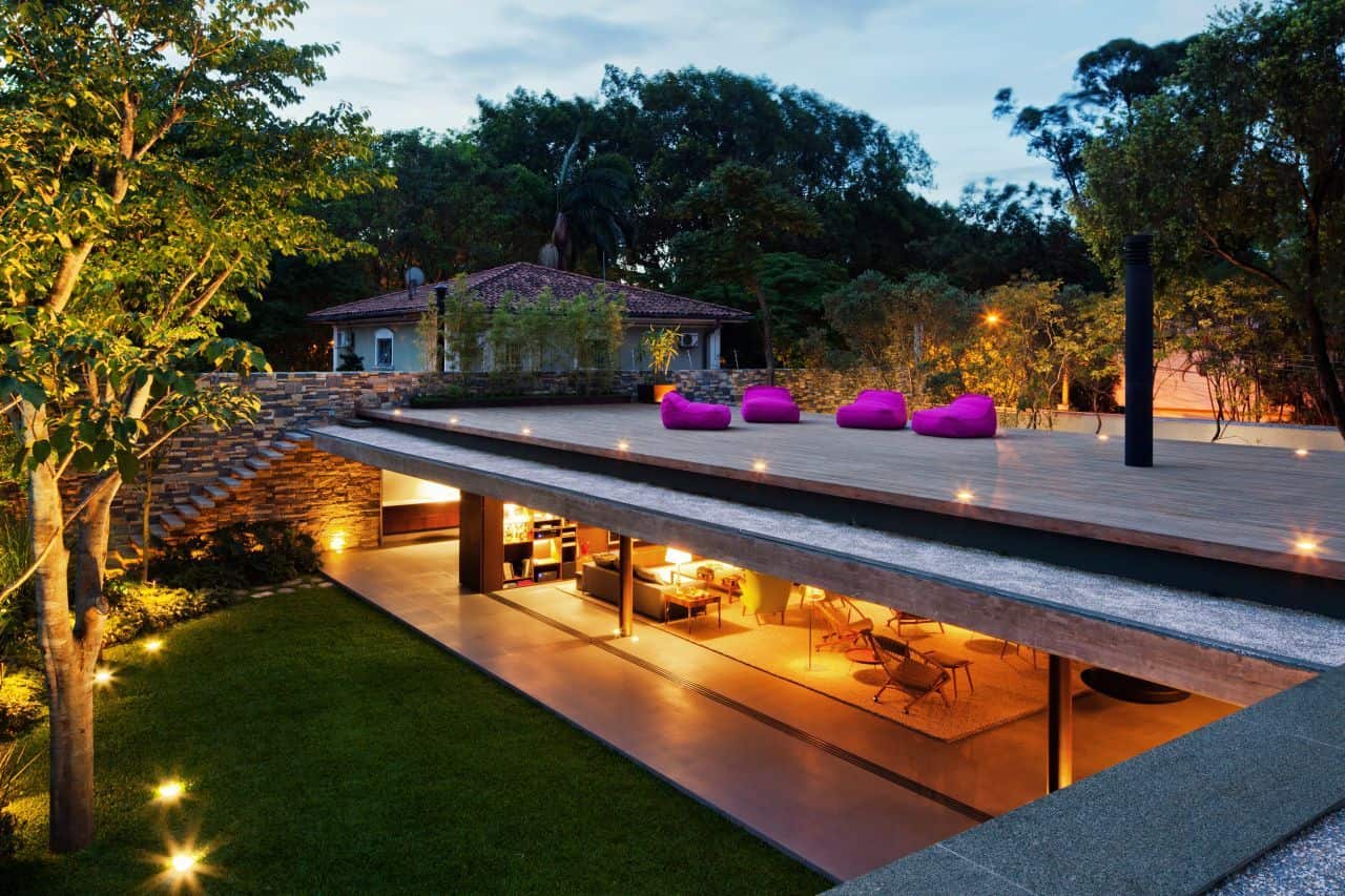 V4 House by Studio MK27: A Ground-Level Pavilion in São Paulo Modern luxury house with illuminated outdoor terrace and vibrant purple lounge chairs, surrounded by lush greenery and trees, showcasing contemporary architectural design.