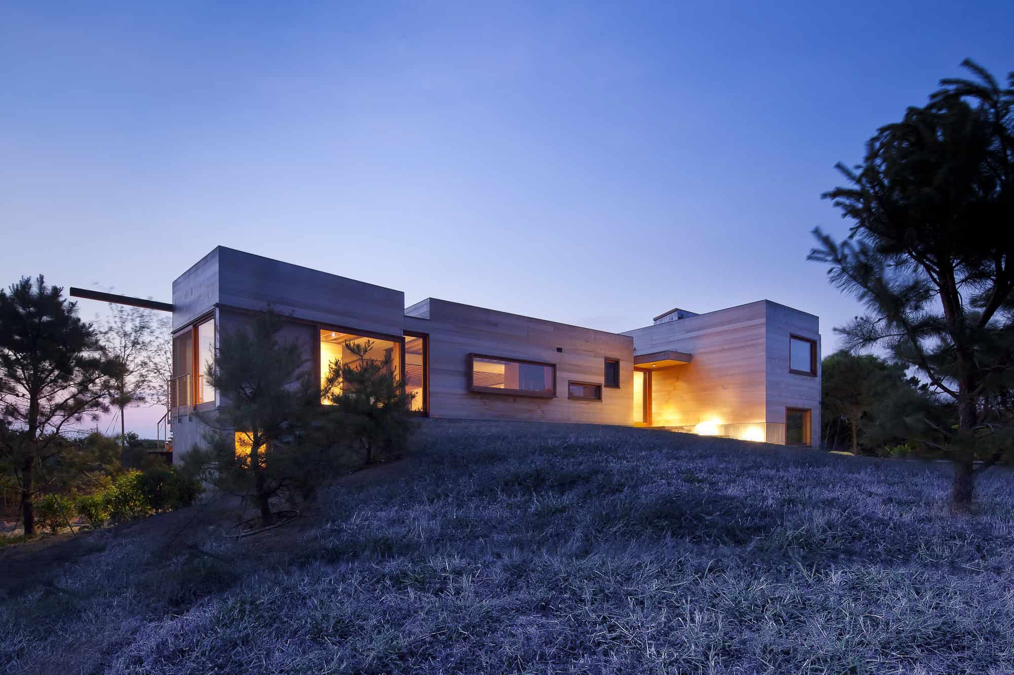 Contemporary modern house with sleek design, large windows, and warm interior lighting, set on a hillside with trees and natural landscape, showcasing innovative architecture and stylish outdoor environment.