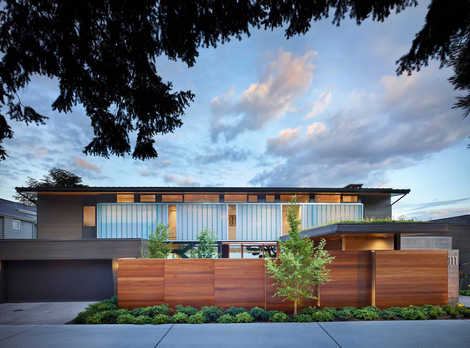 Contemporary modern house with sleek wood siding, large glass windows, and a landscaped front yard under a cloudy sky.