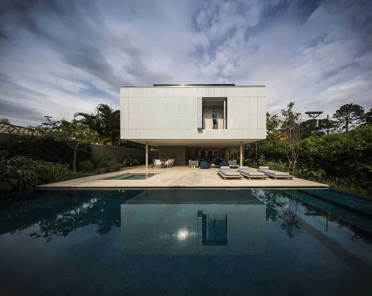 Modern minimalist house with a sleek white facade, elevated structure, and expansive outdoor pool with lounge area, surrounded by lush greenery and designed for contemporary luxury living.