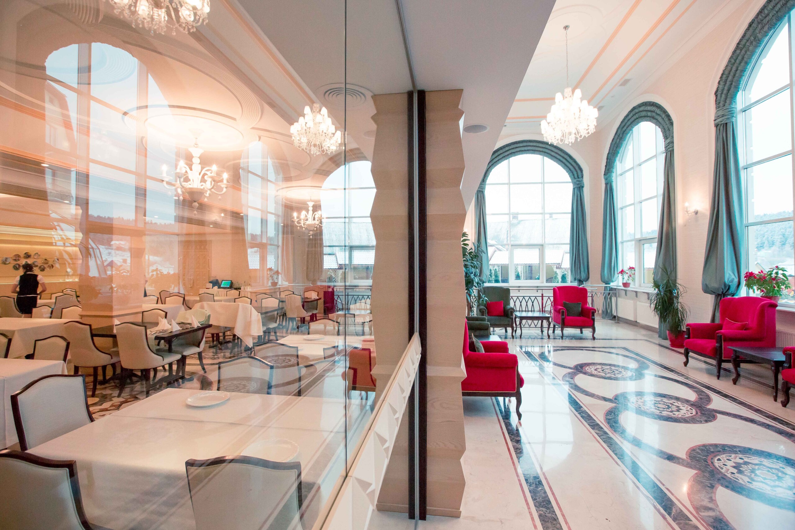 Elegant hotel lounge with large arched windows, crystal chandeliers, and luxurious red and green seating in a classical interior design.
