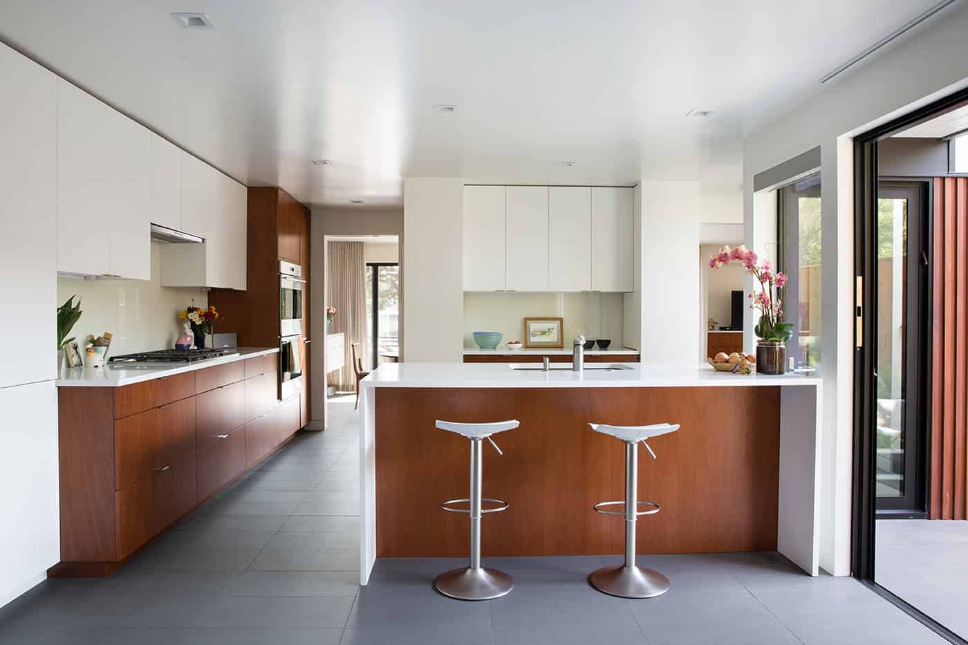 Sleek modern kitchen with minimalist design, wooden cabinetry, white countertops, and open layout, featuring a kitchen island, modern bar stools, and large sliding glass doors, perfect for contemporary architecture art designs.