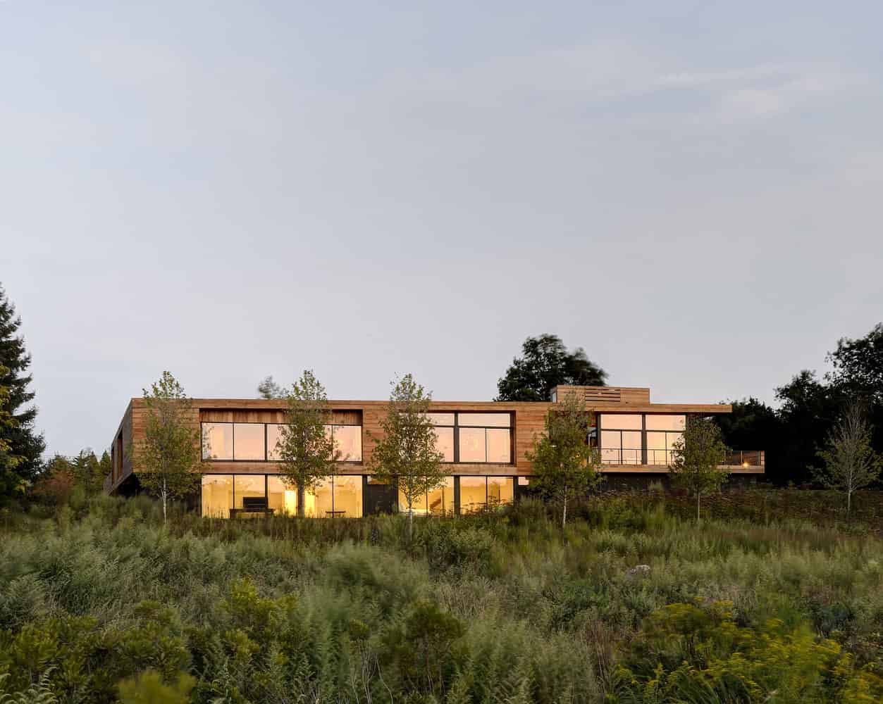 Modern eco-friendly house with large glass windows and wooden exterior, set on a lush green hillside during sunset.