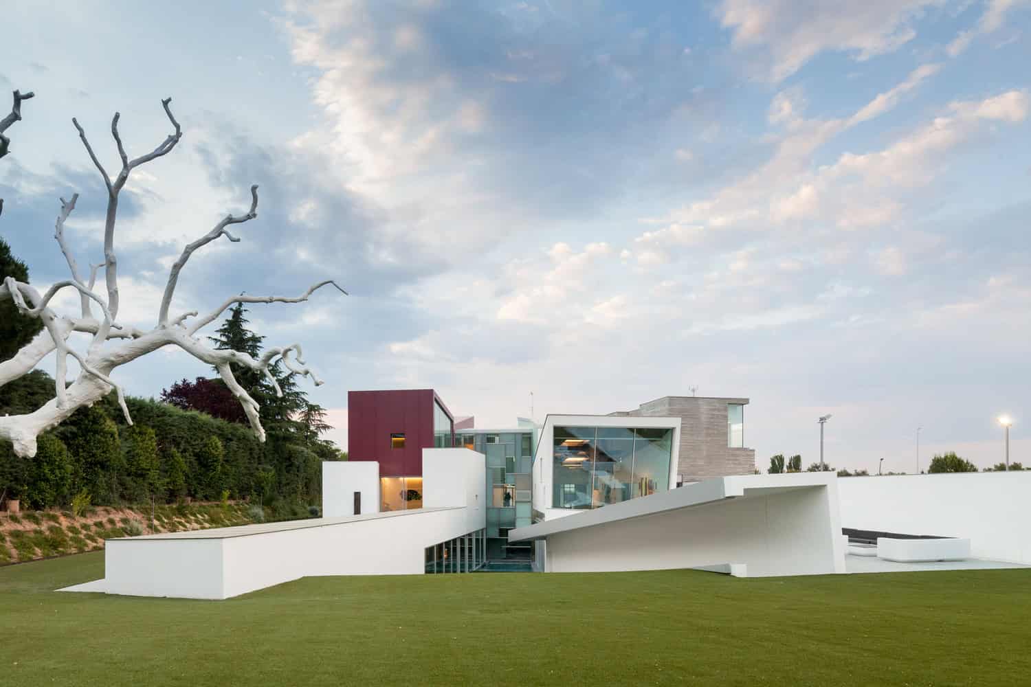 Modern architectural design featuring sleek white structures, expansive glass windows, and innovative shapes, set against a landscaped outdoor area with a dramatic tree sculpture, exemplifying contemporary art in architecture.