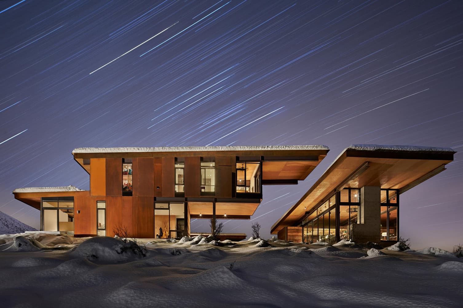 Modern mountain retreat house with contemporary architecture, large glass windows, and unique roof designs surrounded by snow under starry night sky.