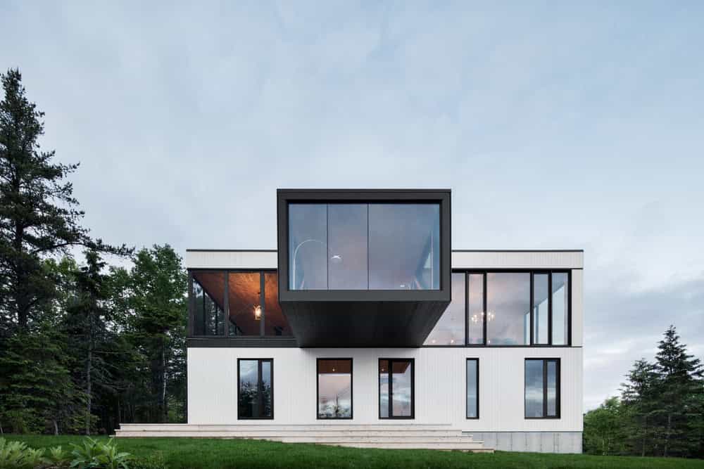 Contemporary modern house with cantilevered upper floor and large glass windows, surrounded by lush greenery and trees, showcasing innovative architecture design.