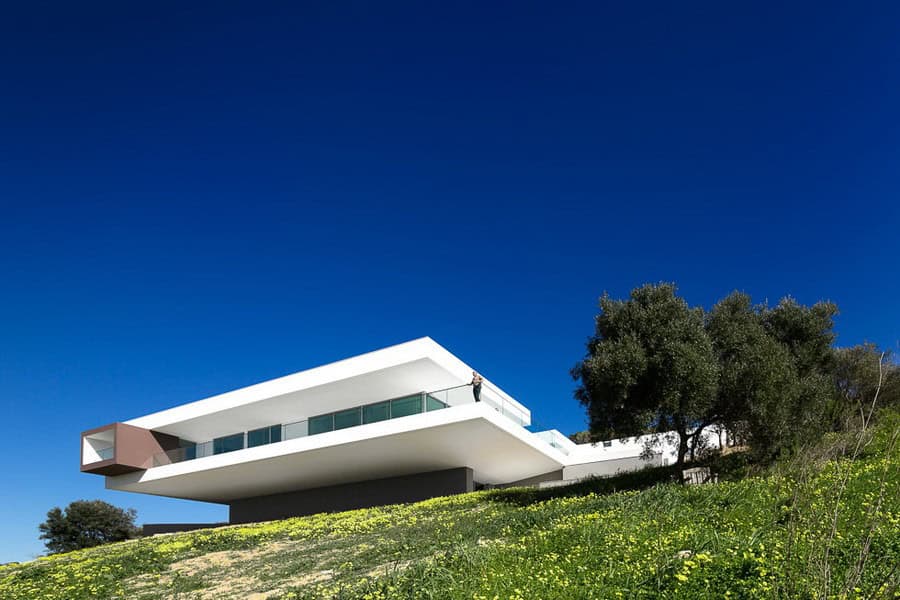 Modern minimalist white house with large glass windows on a hillside under a clear blue sky, showcasing contemporary architecture and innovative design.