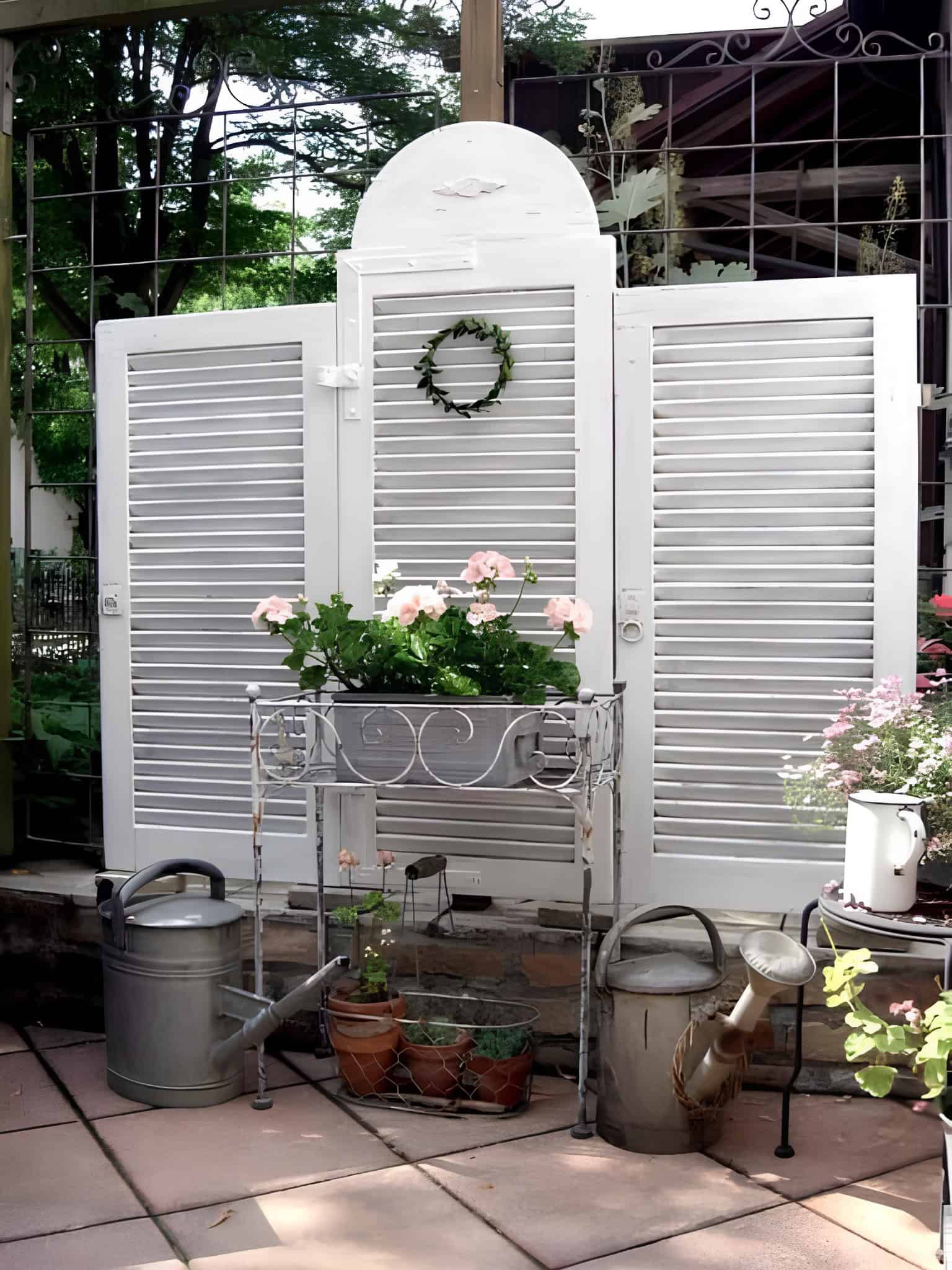 Vintage white shutter panel used as a garden privacy screen with flowers and watering cans