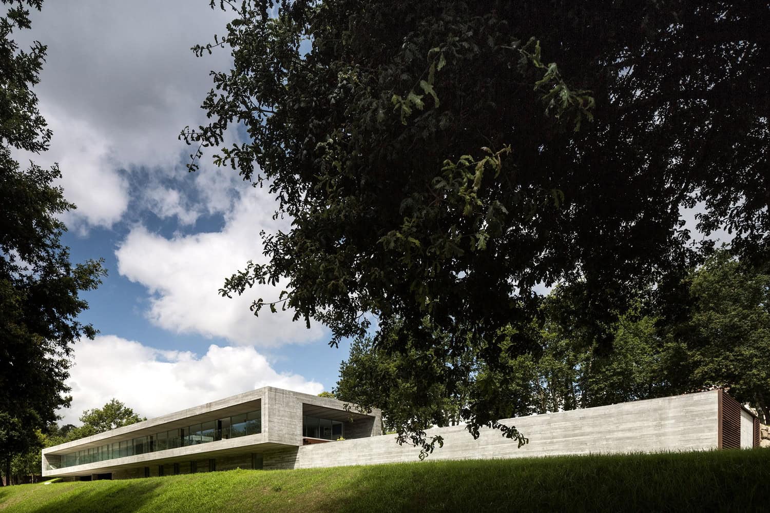 Modern minimalist concrete house with large glass windows, surrounded by lush greenery, under a partly cloudy sky.