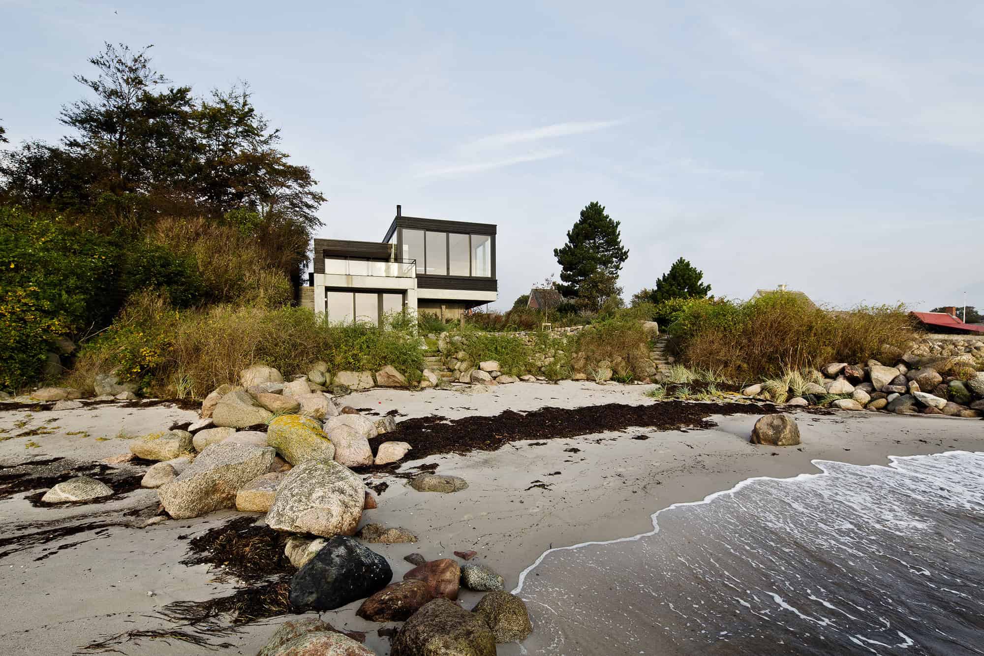 Modern beach house with large glass windows and contemporary architecture overlooking the shoreline, surrounded by coastal landscaping and natural rocks.