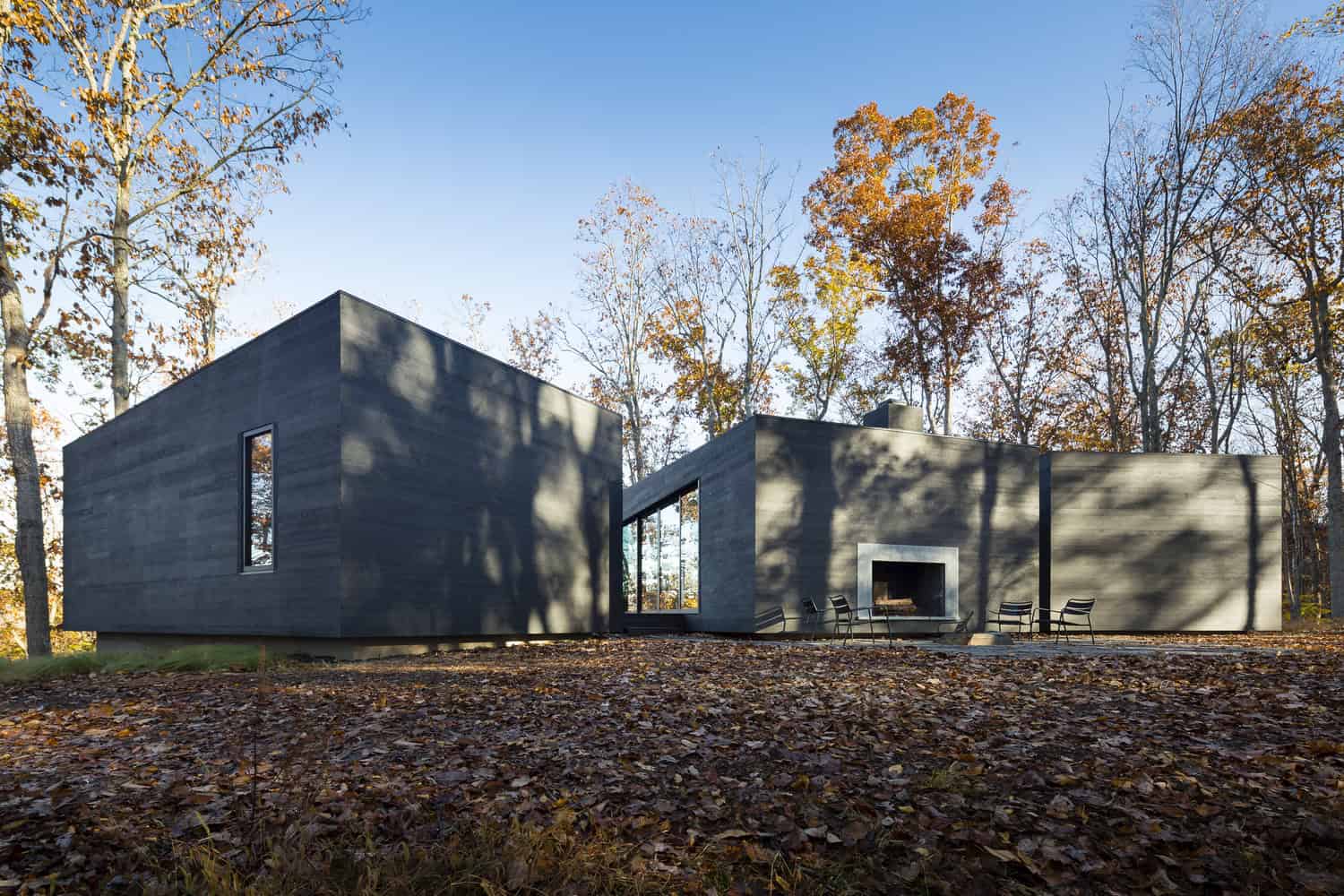 Modern minimalist house with sleek concrete exterior, large windows, and outdoor fireplace surrounded by fall foliage for contemporary architecture and innovative design.