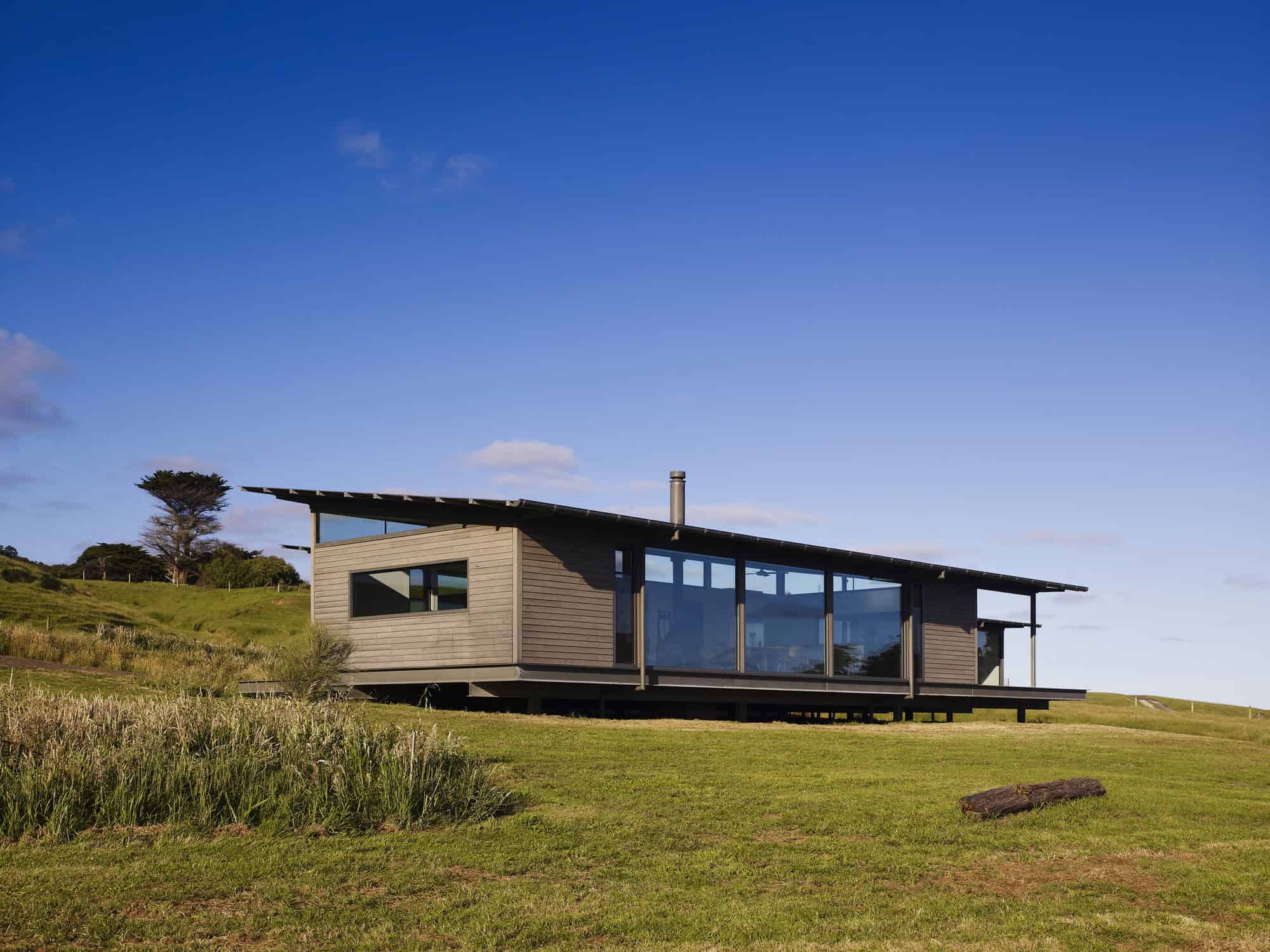 Contemporary modern house with sleek wooden exterior and large glass windows set on scenic rolling green landscape under a bright blue sky.