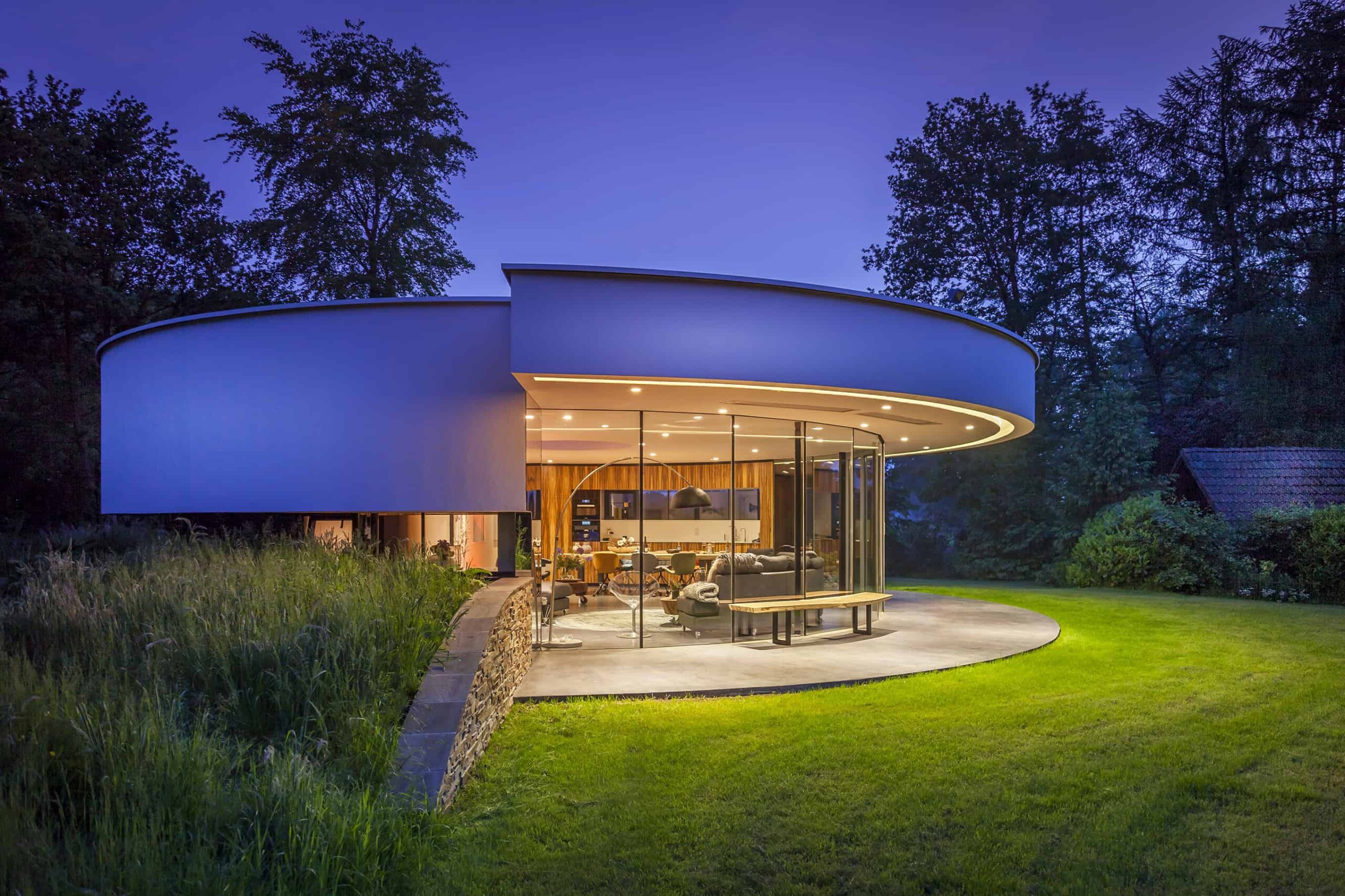 Futuristic modern house with curved architecture and large glass windows, surrounded by lush greenery and trees, illuminated during twilight, showcasing contemporary architectural design and innovative construction techniques.