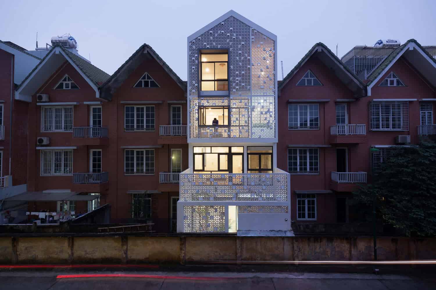 Modern narrow building with intricate lattice design facade illuminated at dusk, surrounded by traditional colorful residential houses, showcasing contemporary architecture and urban design.