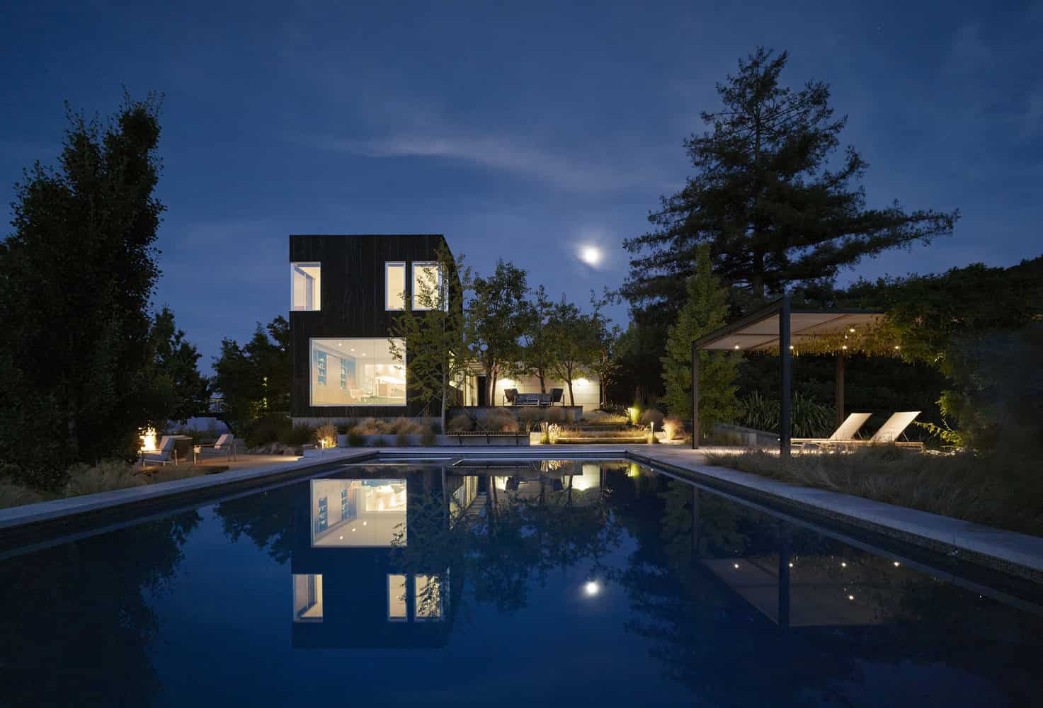 Modern black exterior house with large windows, illuminated outdoor pool and landscaped garden at night under moonlight.