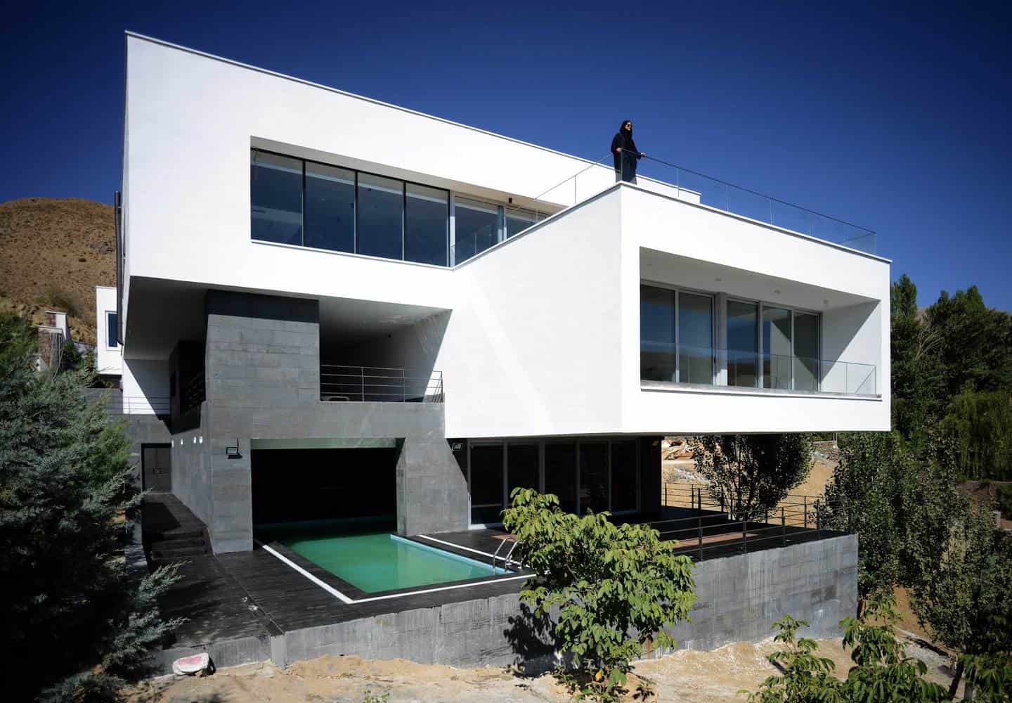 Modern minimalist white house with large glass windows and a rooftop terrace, surrounded by greenery and set on a hillside.