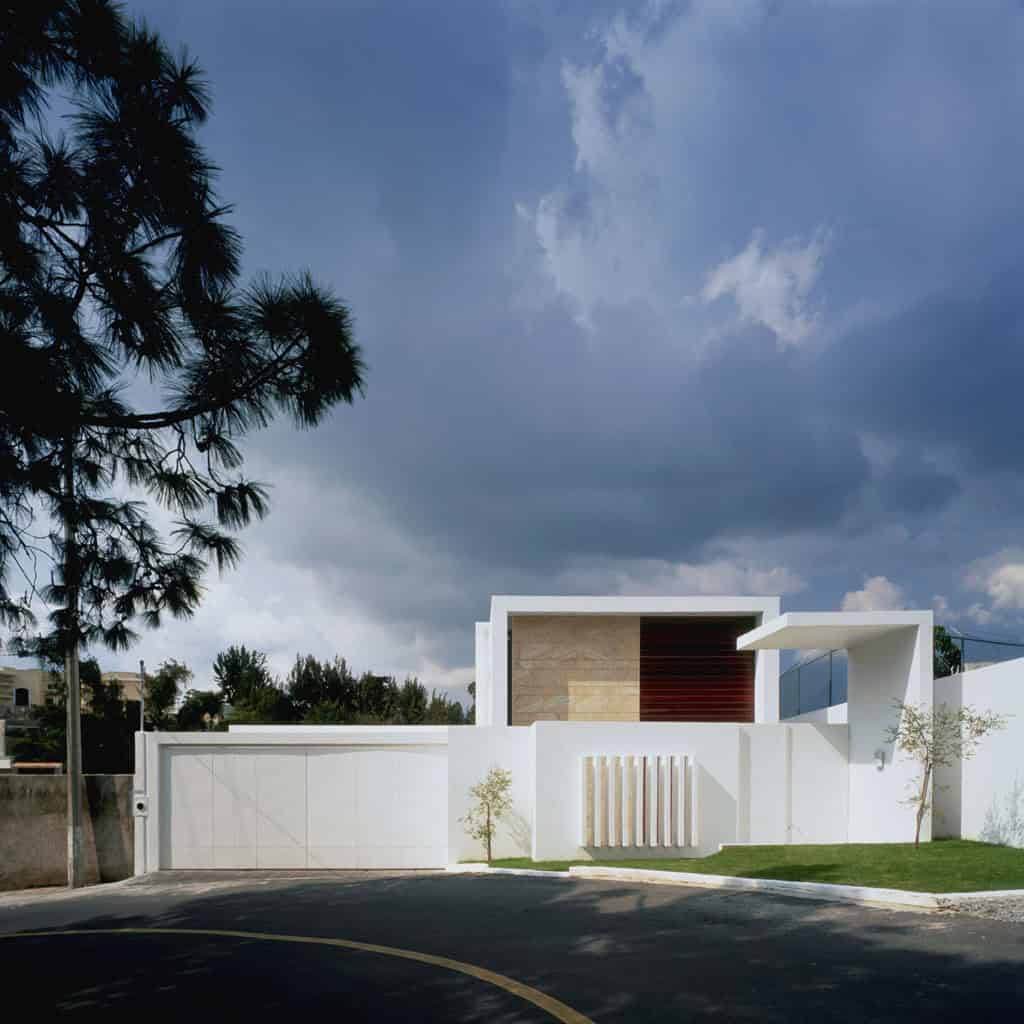 Modern minimalist house with clean lines and white exterior, featuring a flat roof, wooden accents, and a sleek gate, set against a dramatic cloudy sky, exemplifying contemporary architecture and design.