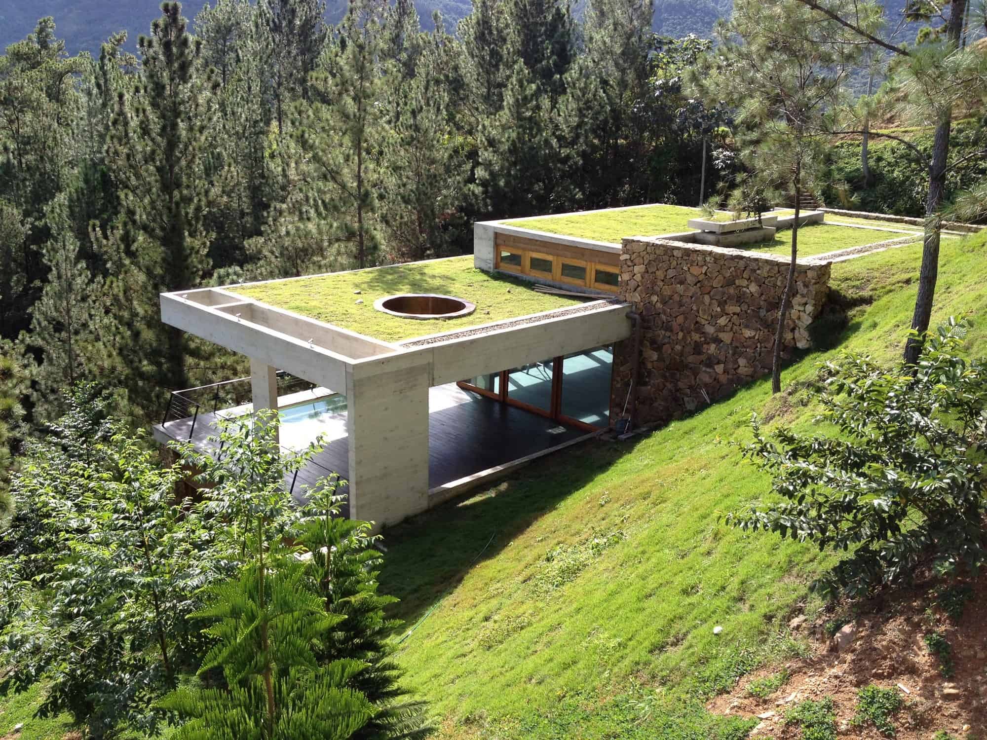 Modern hillside house with flat green roofs, combination of concrete, stone, and glass, surrounded by lush trees, emphasizing innovative architecture and integration with natural landscape.