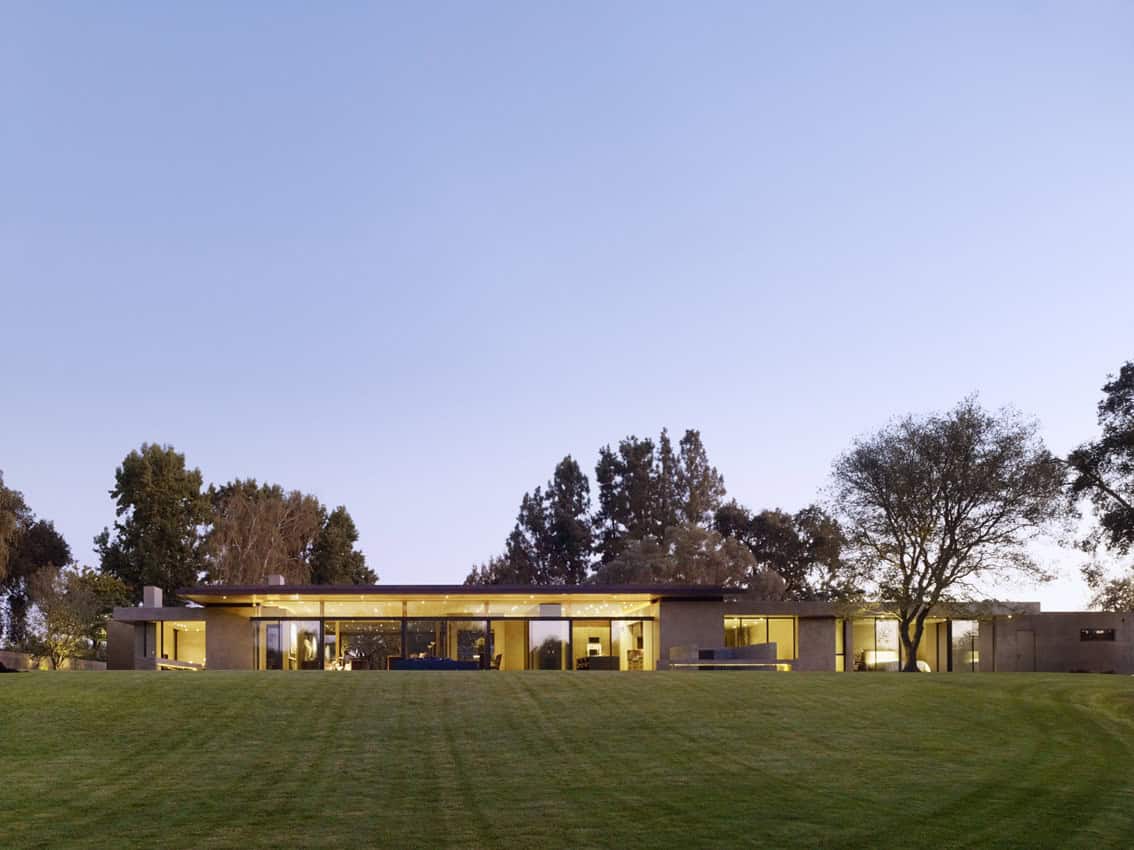 Modern single-story house with large glass windows, surrounded by lush lawn and tall trees, showcasing contemporary architecture and innovative design.