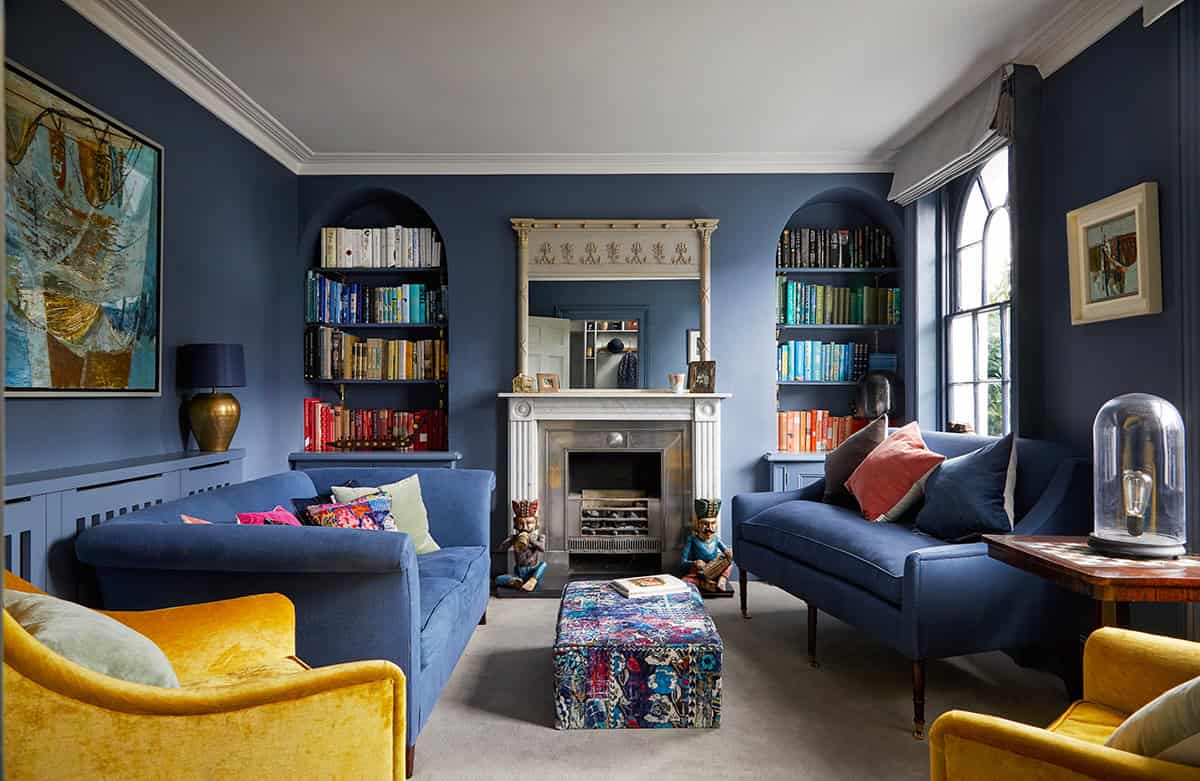 Luxurious living room with navy blue walls, elegant fireplace, colorful velvet sofas, and built-in bookshelves filled with books, creating a cozy and stylish ambiance.