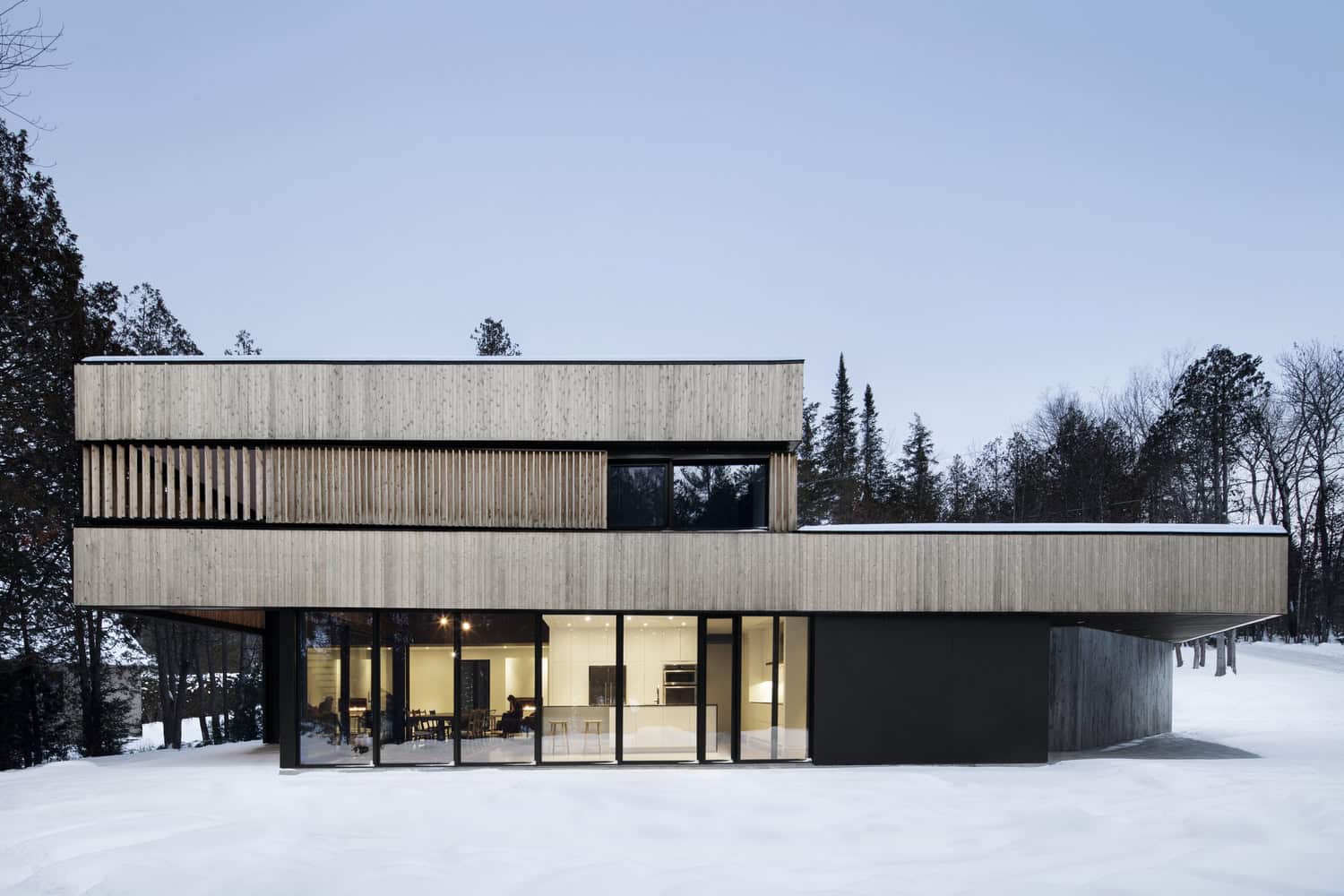 Contemporary modern house with wooden exterior and large glass windows in a winter landscape.