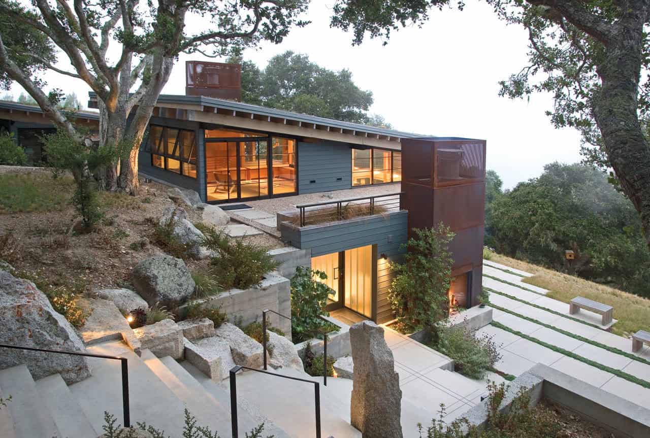 Modern hillside house with large glass windows and contemporary architecture, surrounded by natural landscape and trees, showcasing innovative design and integration with nature.