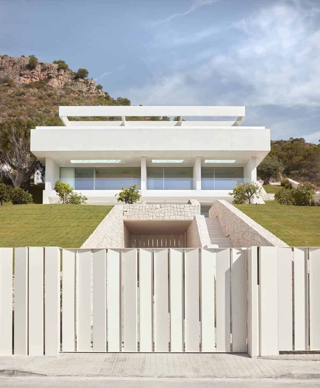 Modern minimalist white house with geometric design, large glass windows, and tiered structure set against a hillside. Features landscaped greenery, stone accents, and a high fence for privacy.
