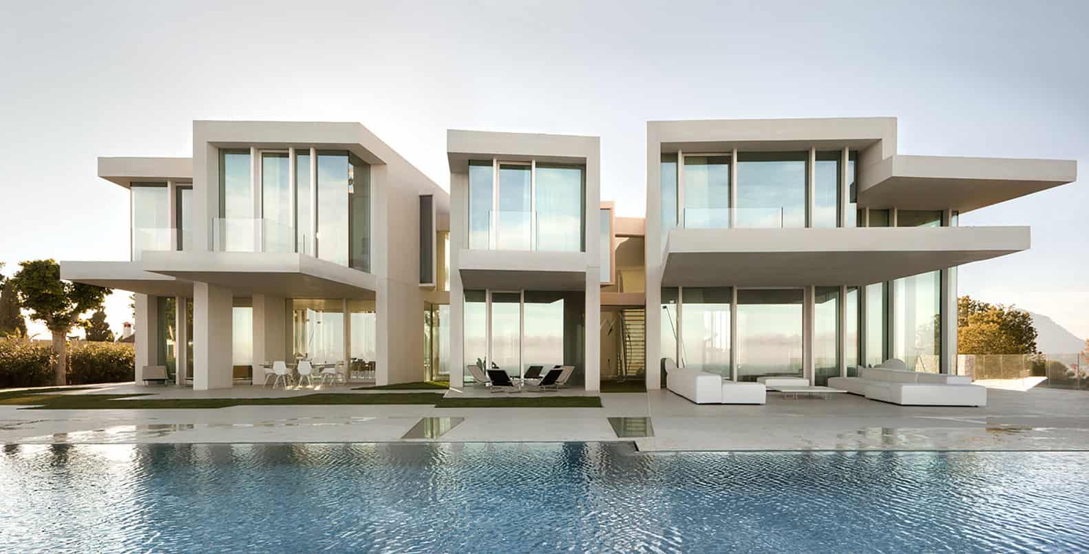 Modern minimalist luxury house with sleek architectural design and large glass windows overlooking a swimming pool in a sunny outdoor setting.