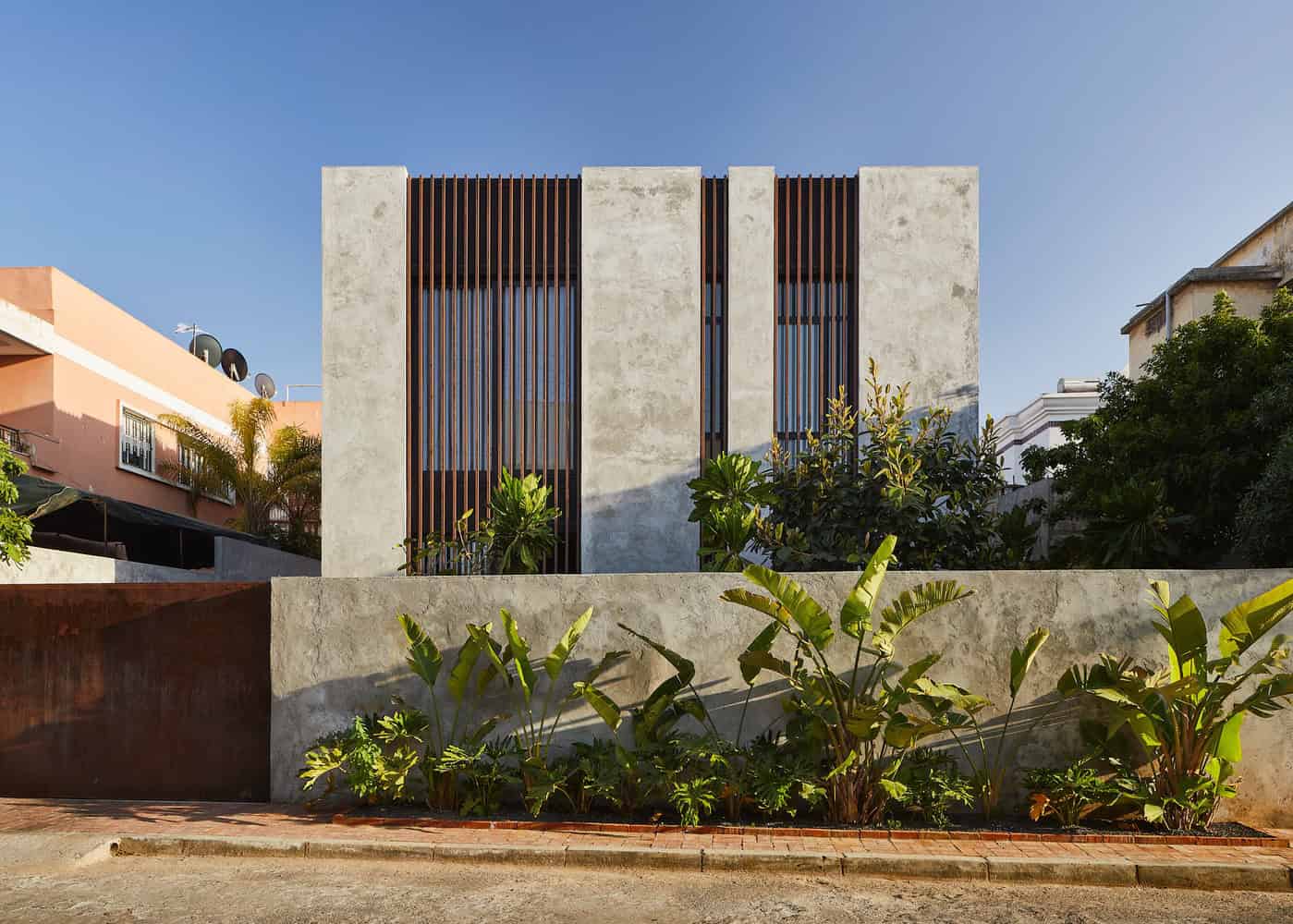 Contemporary modern house with minimalist concrete facade and vertical wooden slats architectural design, surrounded by lush greenery and tropical plants in an urban setting.