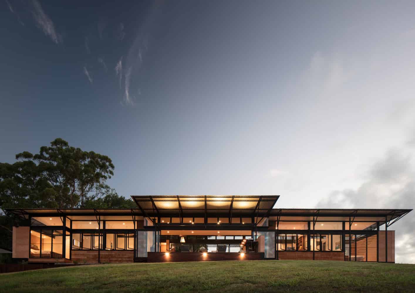 Contemporary modern house with glass walls, metal roof, and open-plan design, set on a grassy landscape during dusk.