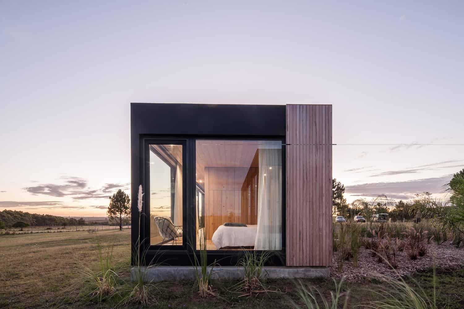 Modern tiny house with sleek black framing, large glass windows, and wooden accents, set in a serene rural landscape showcasing innovative architecture and efficient use of space.