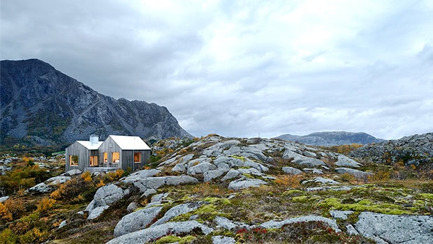Modern minimalist mountain cabin surrounded by rugged natural landscape and rocky terrain with dramatic mountain backdrop.