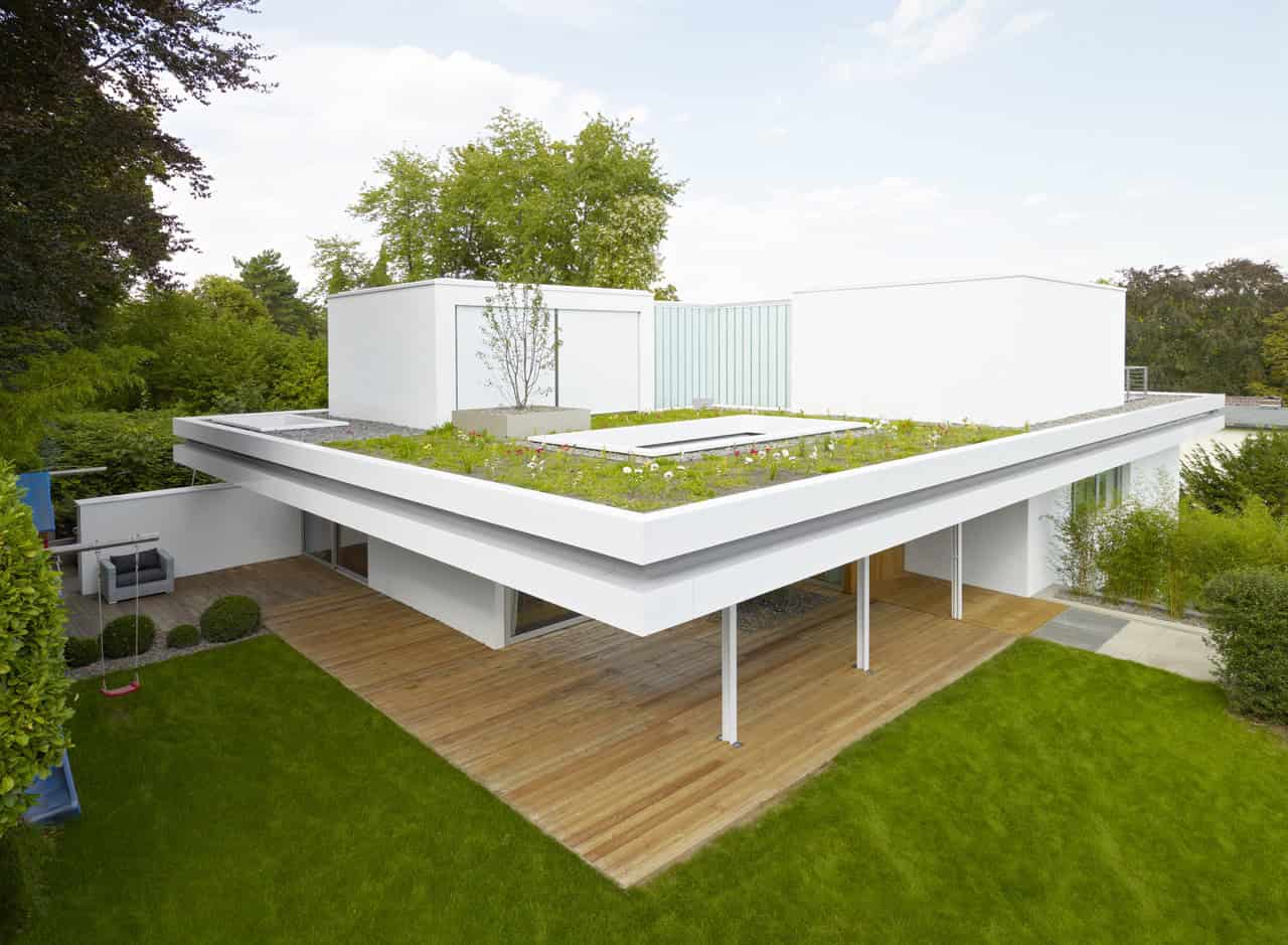 Contemporary modern white house with flat roof and green rooftop garden, surrounded by lush trees and manicured lawn, showcasing innovative architecture and sustainable design.
