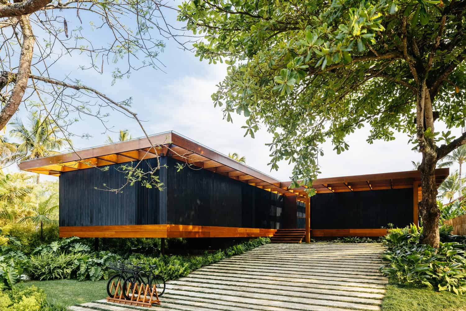 Contemporary black wood house with large overhanging roof, surrounded by lush green tropical garden, modern landscape design, and natural materials for elegant outdoor architecture.