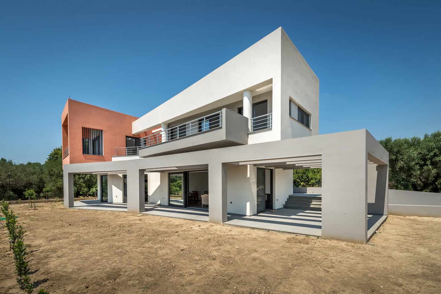 Modern minimalist house with geometric design and clean lines, featuring white and terracotta exterior walls, large glass windows, and spacious outdoor terraces, set in a natural landscape.