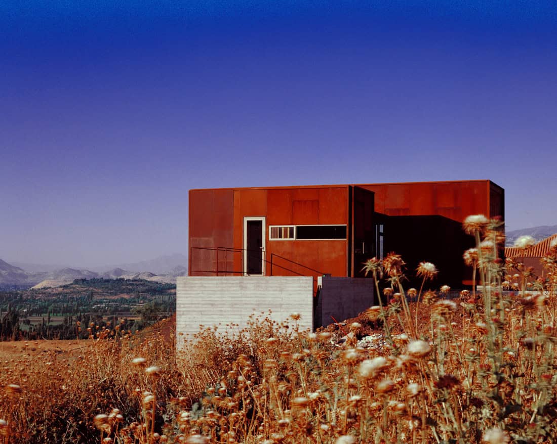 Modern minimalist house with rust-colored metal exterior and concrete accents, set against a mountainous landscape with a vivid blue sky, showcasing innovative architecture designs.