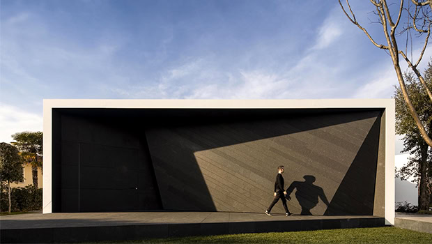 Sleek modern minimalist garage with black textured facade and white frame, featuring geometric design and shadow play, set in a landscaped area with clear blue sky.