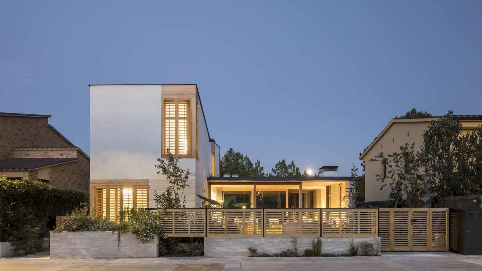 Contemporary modern house with sleek design and large glass windows, illuminated warmly at dusk, surrounded by a wooden fence and lush greenery.