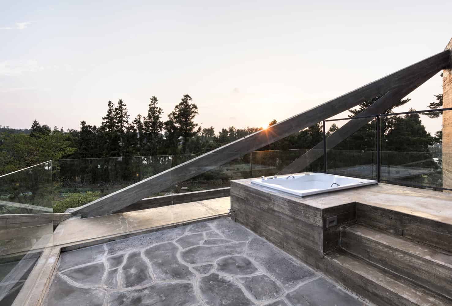 Modern rooftop balcony with glass railing, outdoor Jacuzzi, and stunning sunset view surrounded by lush trees, showcasing contemporary architecture and stylish outdoor living spaces.