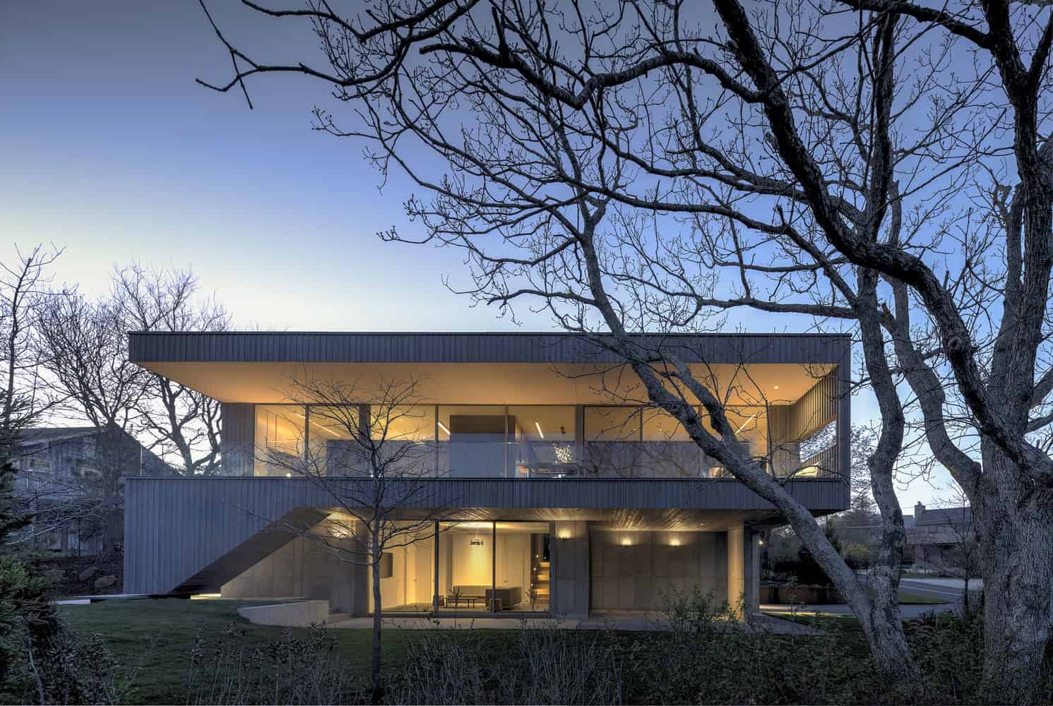 Modern minimalist house with sleek architectural design, large glass windows, and clean lines, surrounded by leafless trees during dusk.