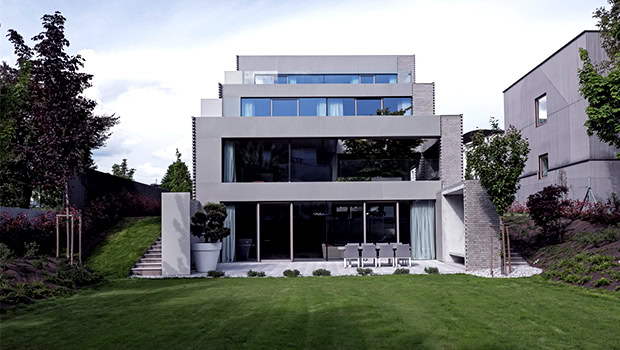 Modern minimalist concrete residential building with large glass windows and tiered terraces, surrounded by lush landscaping and green lawn.