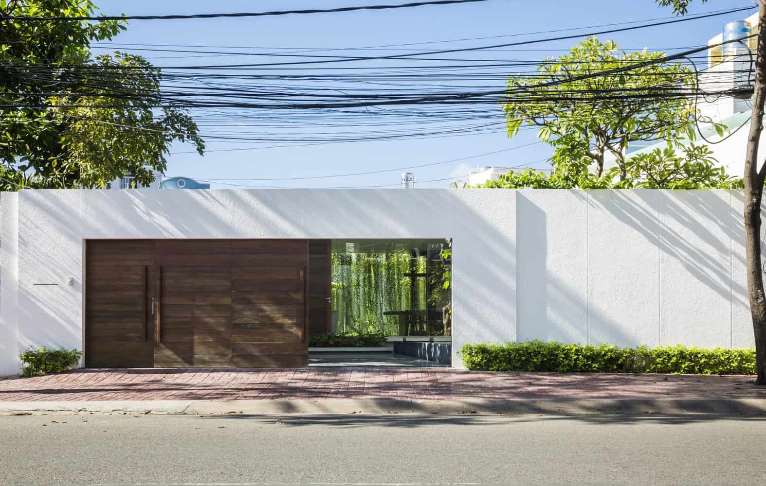 Contemporary modern house exterior with clean white wall, wooden gate, lush greenery, and overhead power lines in a minimalist design.